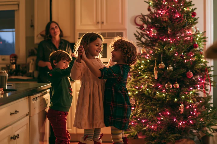 Three children dance beside a decorated Christmas tree in a warmly lit home.