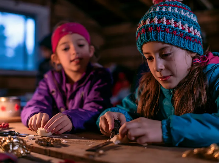Two children sit at a wooden table inside a workshop, focused on making handmade ornaments.