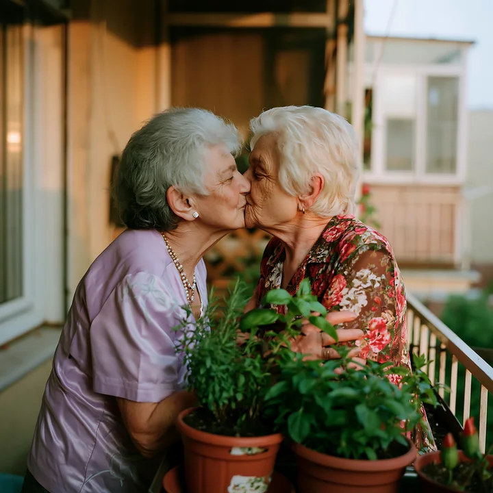 Two elderly partners kiss on a porch surrounded by leafy potted plants.