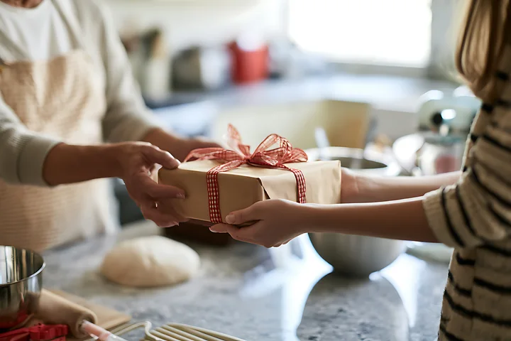 Two hands are exchanging a wrapped gift in a kitchen while preparing for a holiday celebration.