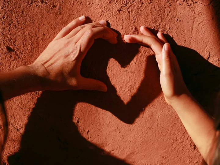 Two hands form a heart-shaped shadow in strong sunlight on a textured terracotta wall.
