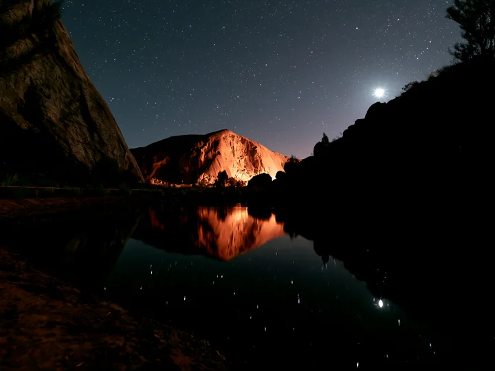 Uluru stands tall under a starry night sky, surrounded by rocky formations.