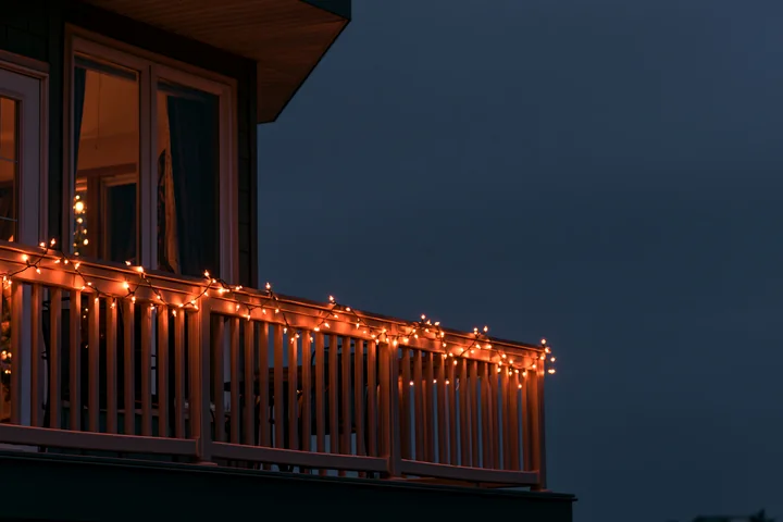 Warm Christmas lights glow along a residential balcony railing at dusk beneath a dark blue sky.