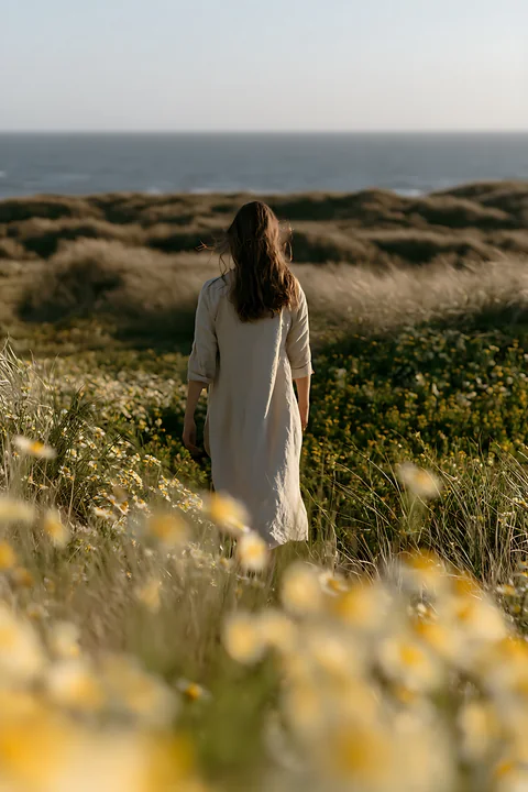 Woman in a white dress walks through sunlit coastal flowers toward the ocean.