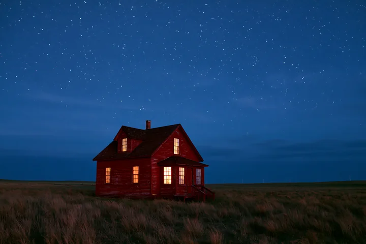 A bright red cabin stands alone in a grassy field, illuminated from within by warm light, under a clear night sky filled with stars.