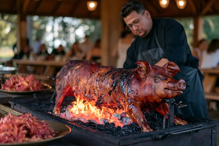 A chef tends to a roast suckling pig over a charcoal grill at an outdoor barbecue catering event.