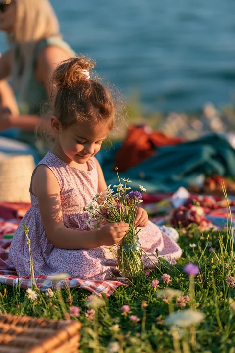 A child sits on a blanket by an alpine lake, carefully arranging wildflowers in a small jar.