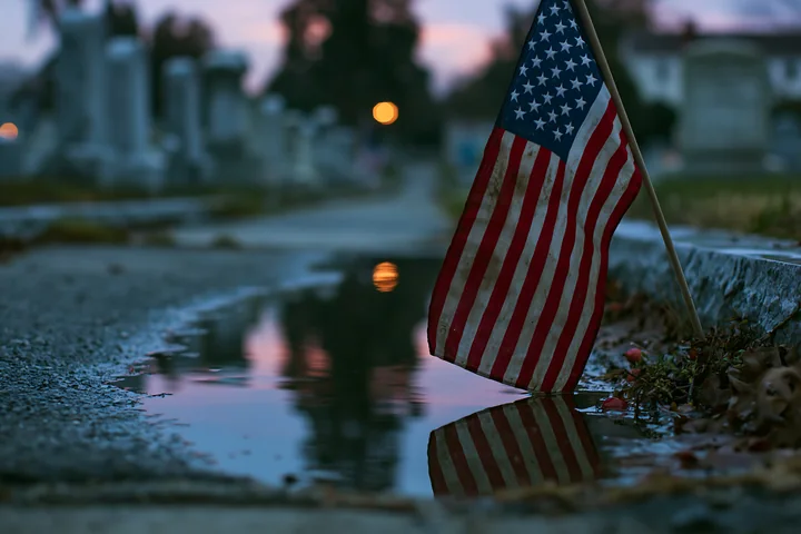 A close-up view of a small American flag at a veterans cemetery, reflecting in a puddle on a pathway as dusk settles over the gravestones in the background.