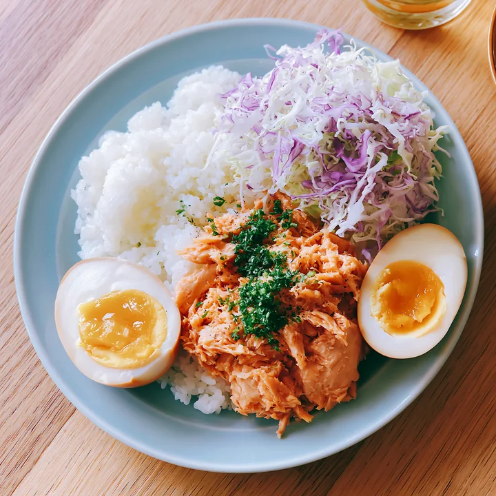 A colorful breakfast plate features white rice, shredded cabbage, marinated fish, and soft-boiled eggs on a wooden table.