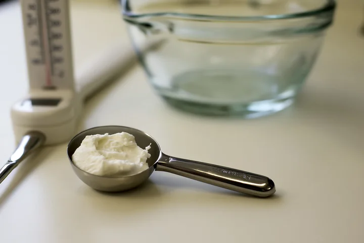 A cosmetic scoop holds a measured dose of cream next to a clear glass bowl and various tools in a well-lit workspace.