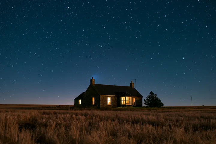 A country house stands alone in a field, illuminated by warm light from its windows.