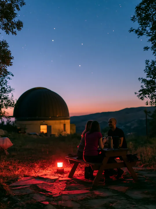 A couple at a remote observatory at night sits at a table under the stars, sharing drinks.