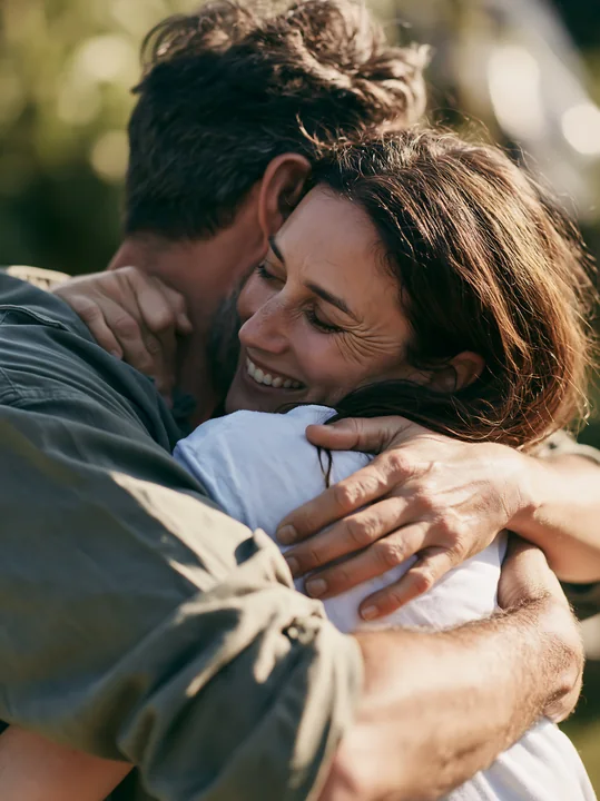 A couple embraces outdoors in spring with smiles, surrounded by green foliage.