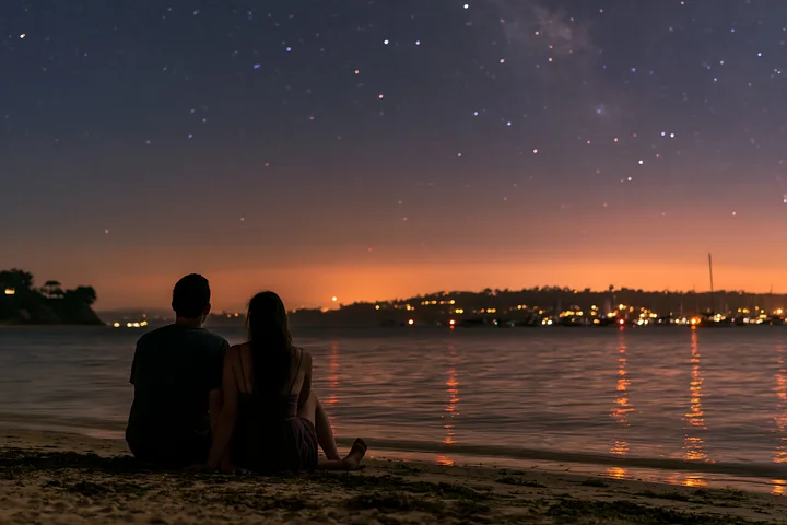 A couple sits together on the beach under stars, watching the night sky.