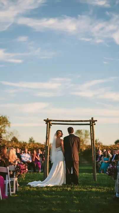 A couple stands closely under a wedding arch during an outdoor ceremony, surrounded by guests seated on chairs.