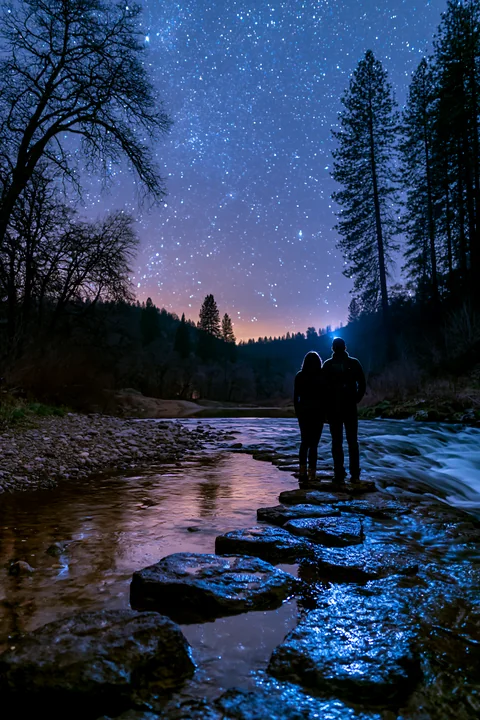 A couple stands on stones by a waterfall, gazing at stars in a clear night sky.