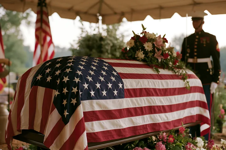 A flag-draped coffin during veteran funeral service stands under a tent surrounded by flowers.