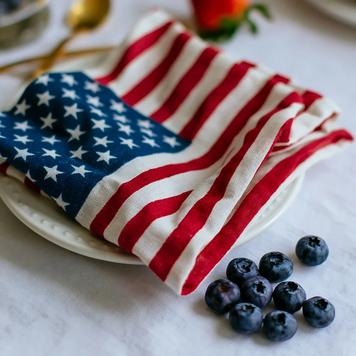 A folded American flag table decoration rests on a white plate next to fresh blueberries.