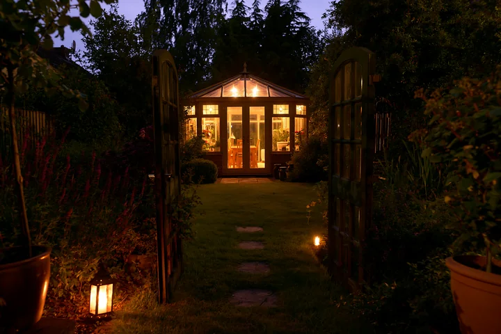 A garden conservatory stands lit from within, surrounded by plants and trees.