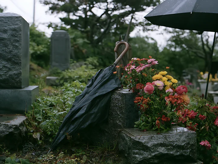 A grave with fresh flowers sits next to an open umbrella in a cemetery during rainy weather.
