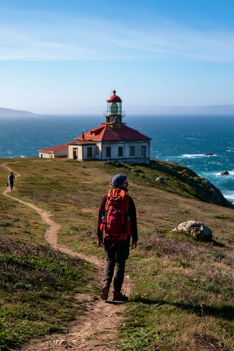 A hiker walks along a dirt path toward an abandoned lighthouse on a remote coast.