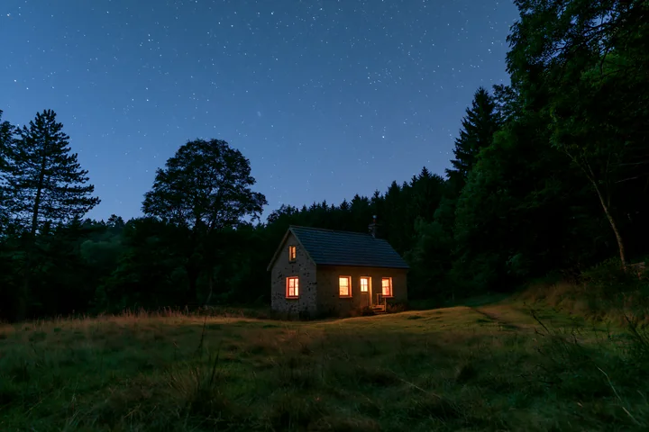 A house sits in a meadow surrounded by trees under a starry sky.