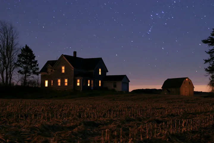 A house under starry night stands in a field, illuminated by warm light from its windows.