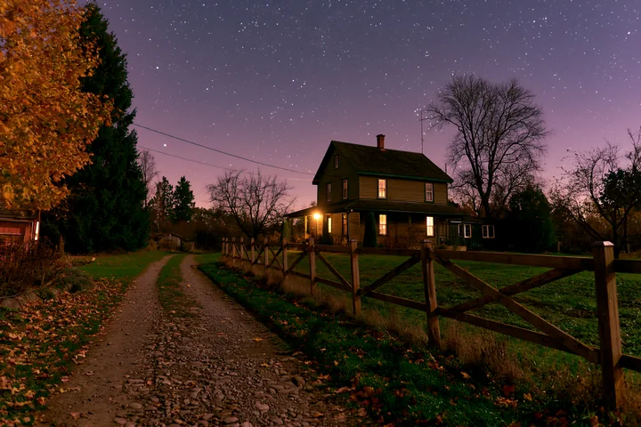 A house under stars with gravel driveway is lit softly by lights.