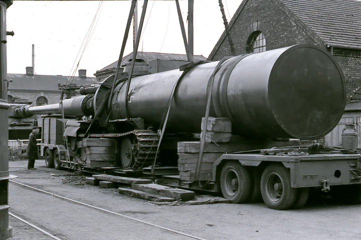 A large piece of artillery is being loaded onto a transport vehicle at a railway yard.
