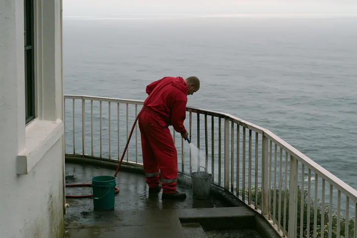 A lighthouse maintenance worker at work cleans the railing and surrounding area near the water.