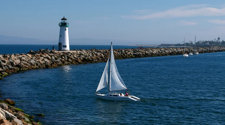 A lighthouse stands tall at the end of a rocky pier, while sailboats glide through the calm waters.