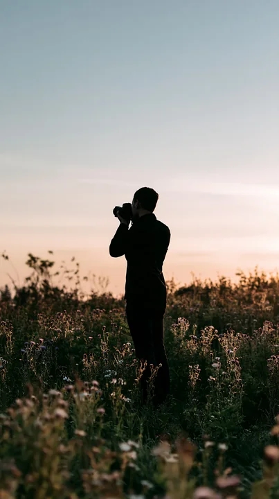 A man is photographing meadow flowers at sunset, standing among wild blooms and tall grass.