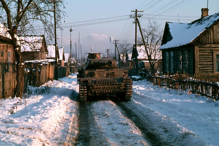 A panzer tank in winter snow drives down a narrow road lined with wooden houses.