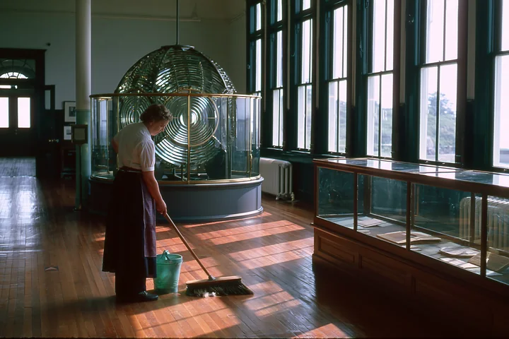 A person cleans the wooden floor in a lighthouse museum interior featuring a large lens display and exhibit cases.