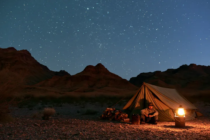 A person sits by a lit lantern at a desert campsite with a tent beneath a sky full of stars.