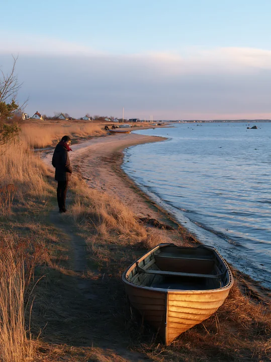 A person stands on a sandy coastline, looking at the water with a small boat nearby.