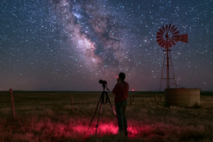 A person using a camera under the Milky Way stands next to a windmill in a rural field at night.