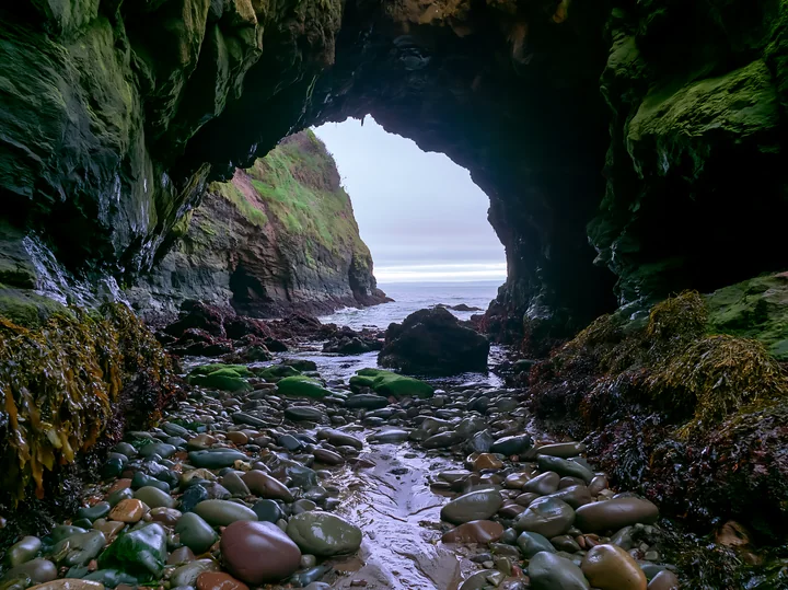 A rocky cave opening reveals a view of the coastline beneath dramatic cliffs.