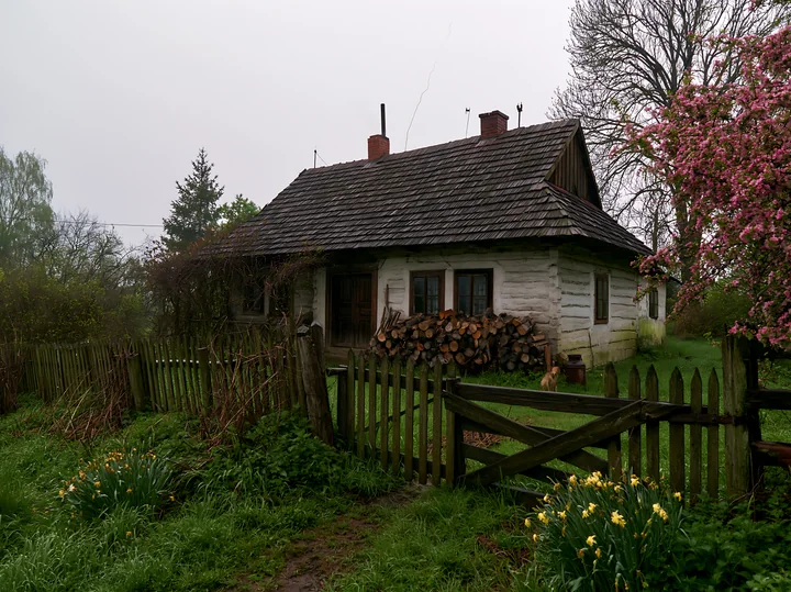 A rural cottage with chimney smoke rises from the roof amid lush greenery and flowering plants.