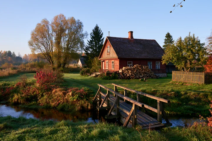 A rural cottage with firewood stack is surrounded by lush greenery and a small stream.