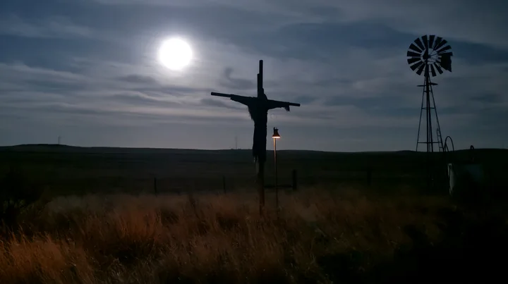 A scarecrow in a moonlit field holds its position against the night sky, while a windmill stands quietly nearby.