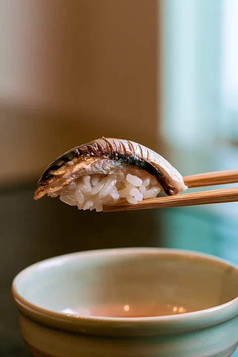 A single sushi piece ready to eat with chopsticks is held above a small bowl.
