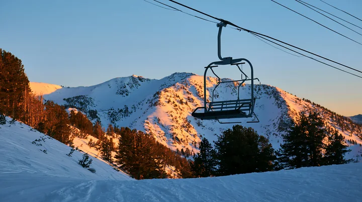 A ski lift stands still in a snowy landscape as the sun sets, casting golden light on the surrounding mountains.