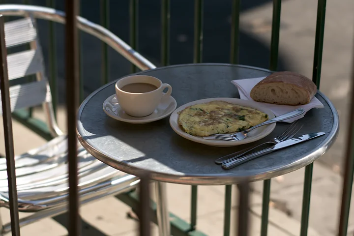 A small cafe table holds a cup of milk coffee, a plate with an omelet, and a piece of bread.