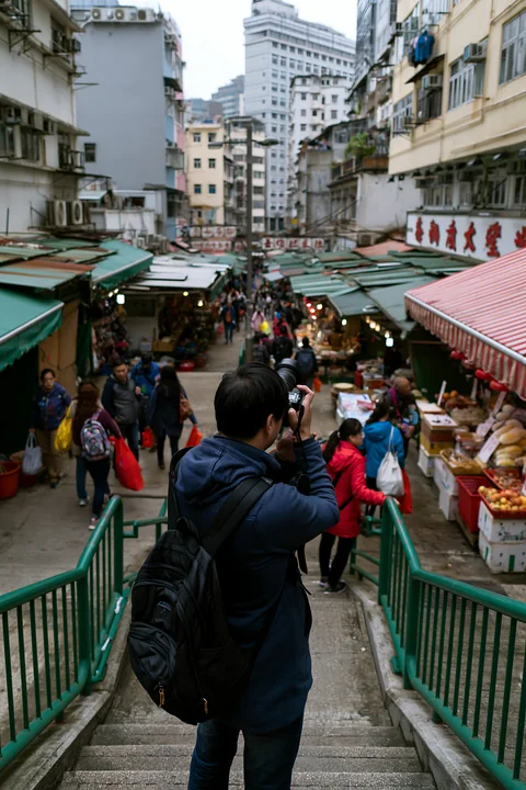 A street photographer is taking pictures in an urban market filled with various stalls and shoppers.