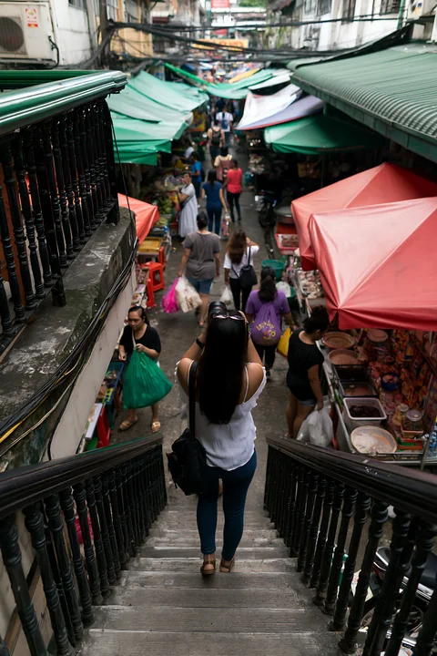 A street photographer taking pictures in an urban market walks down the stairs onto a crowded street filled with vendors and shoppers.