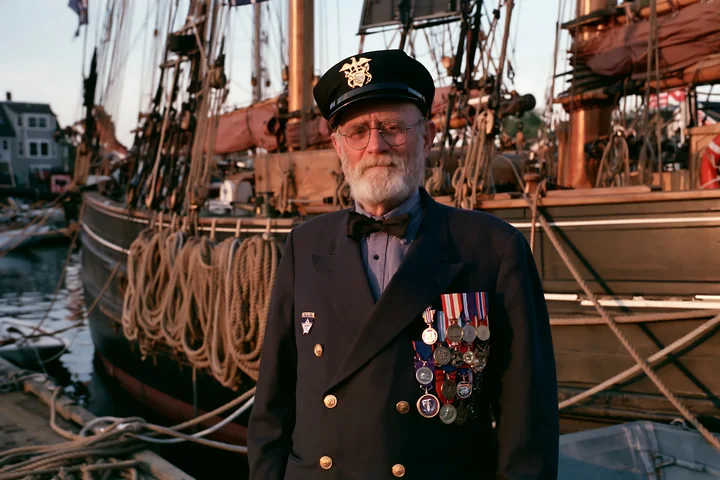 A veteran wearing medals poses near a historic wooden ship in the harbor during golden hour.