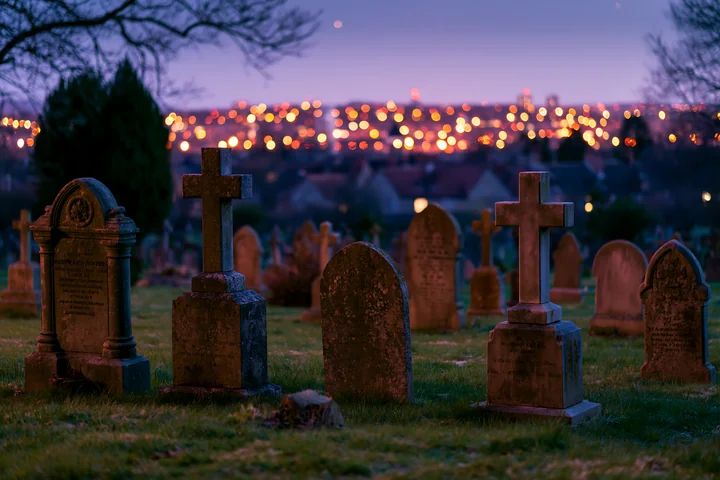 A war cemetery features headstones and crosses, with distant city lights glowing under a starry sky.