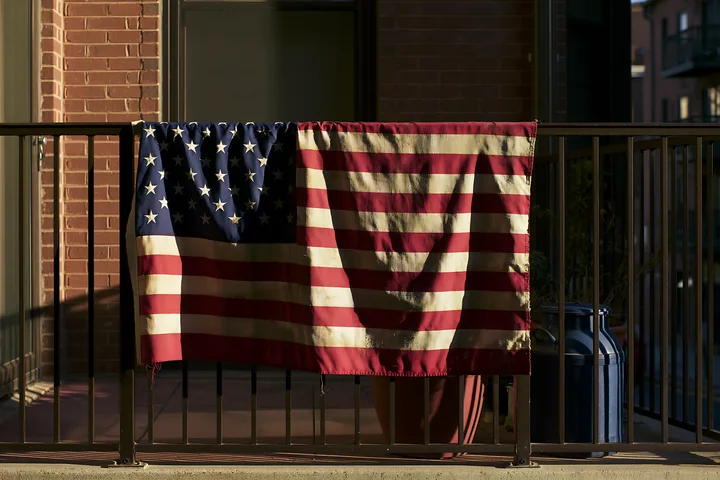 A weathered American flag drapes over a balcony railing, showing signs of fading and wear.
