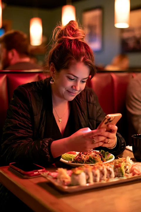 A woman enjoys a sushi dinner under warm restaurant lighting while looking at her phone.